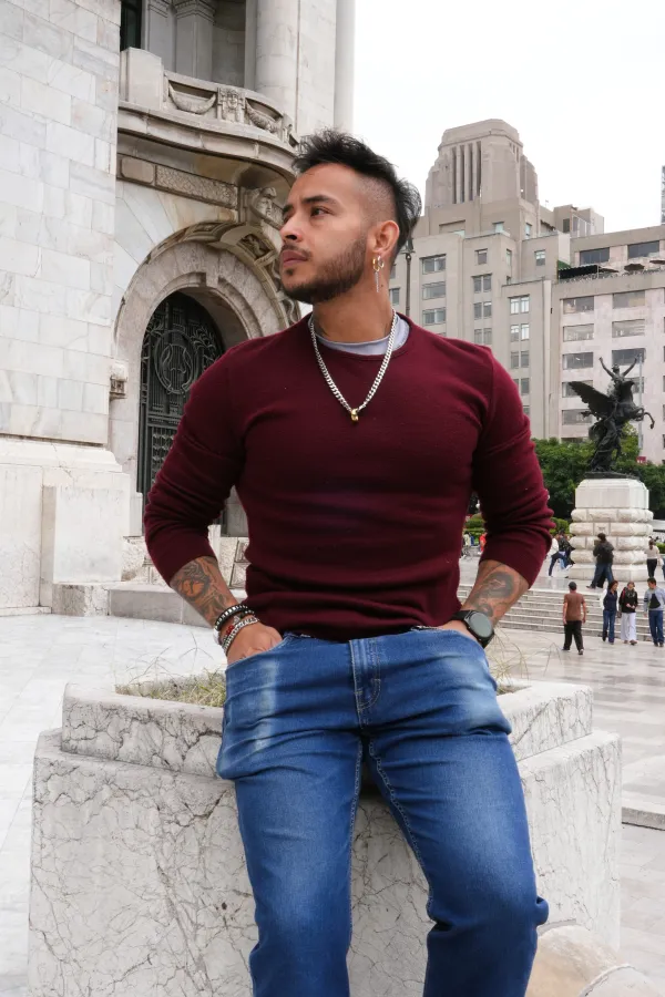 Hombre con suéter guinda y jeans recargado en un muro frente a la fachada del Palacio de Bellas Artes